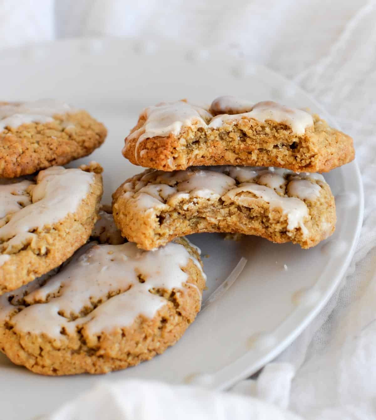 Several iced oatmeal sourdough cookies on a white plate, with one cookie broken in half to show its soft, chewy interior.