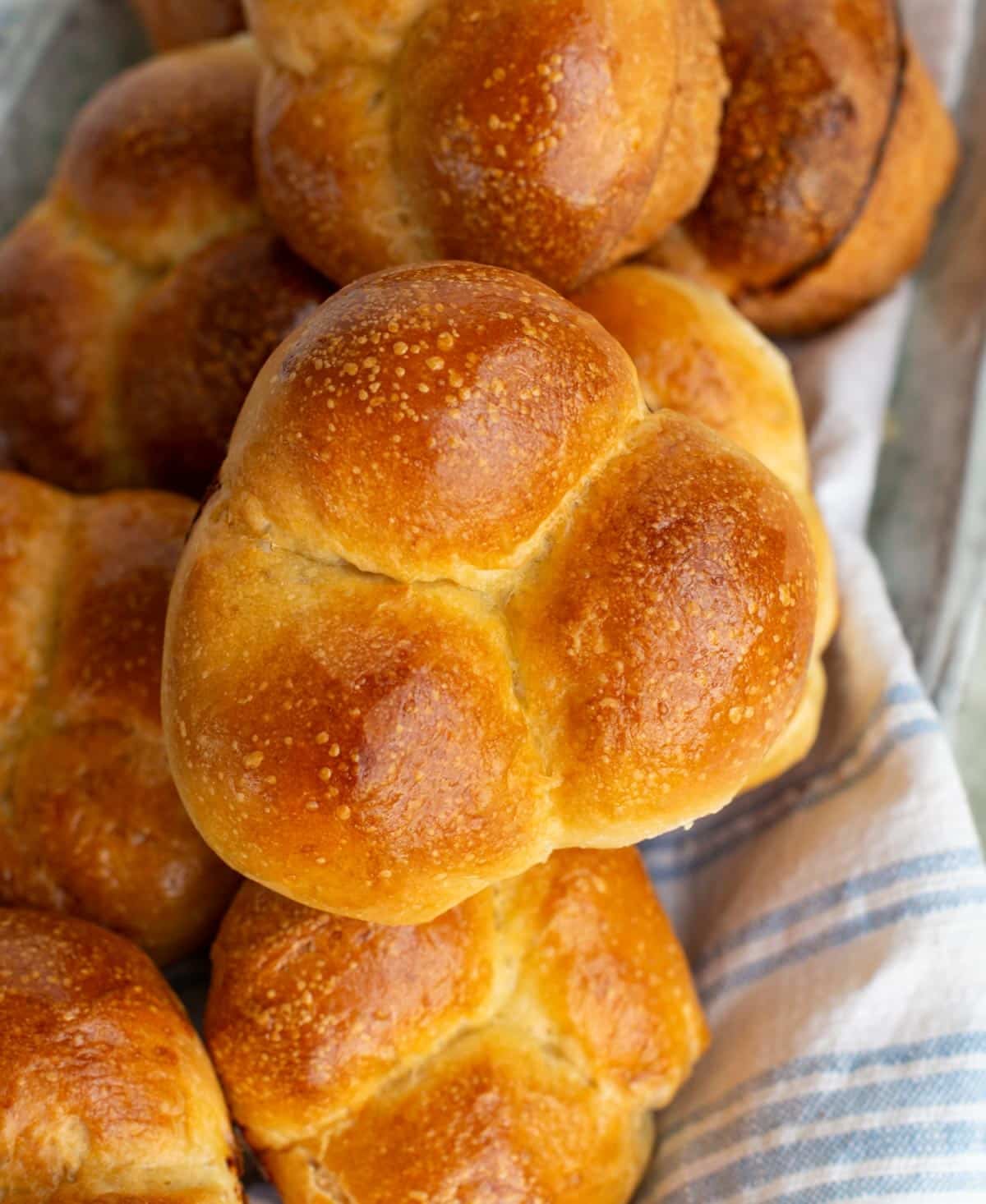 A close up shows brown Cloverleaf Sourdough Rolls arranged in a basket lined with a white and blue striped kitchen towel, showing their soft, fluffy texture and signature three-ball clover shape.