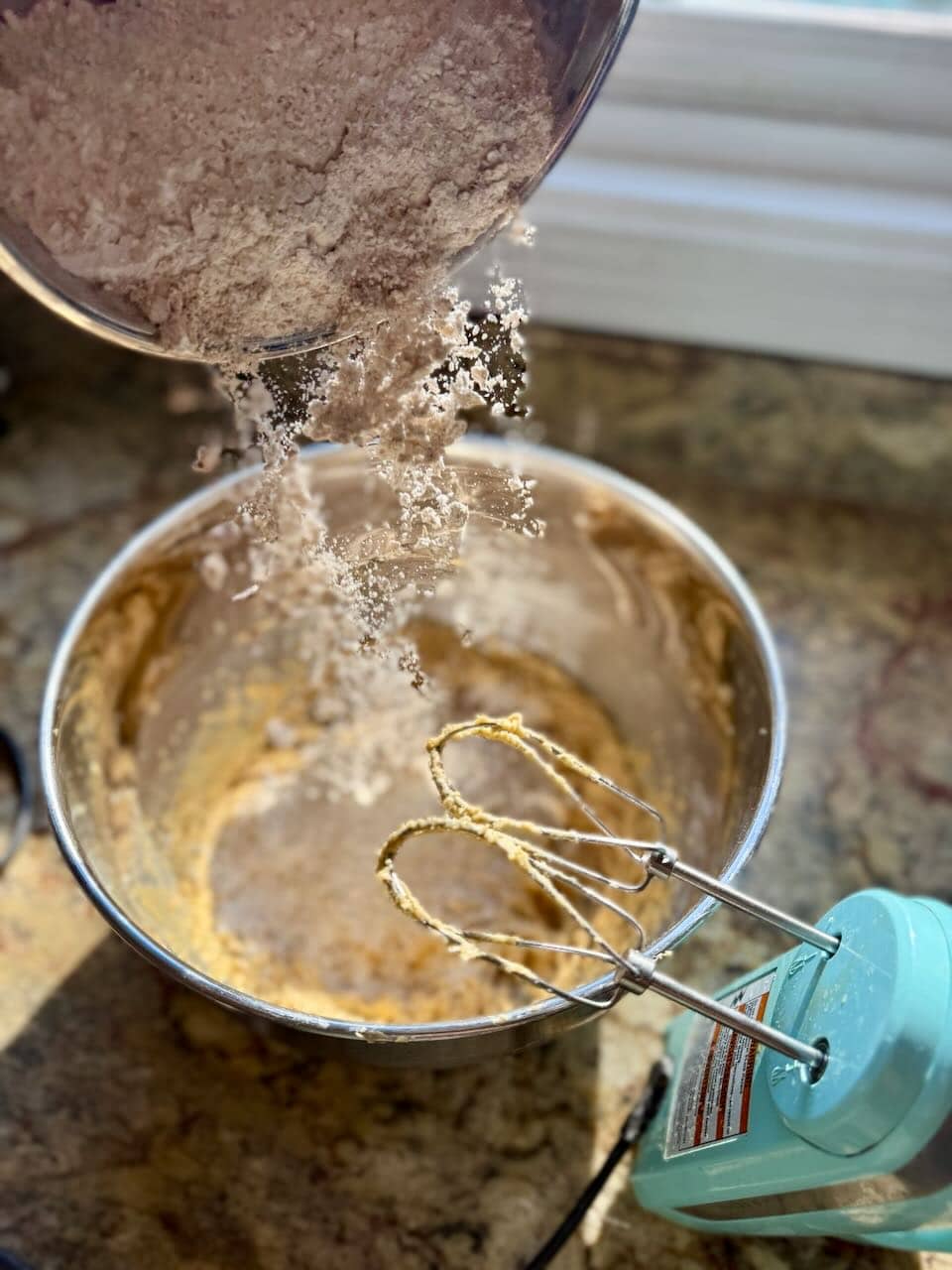 Dry ingredients being poured into the wet mixture in a metal bowl, with a hand mixer ready to blend.
