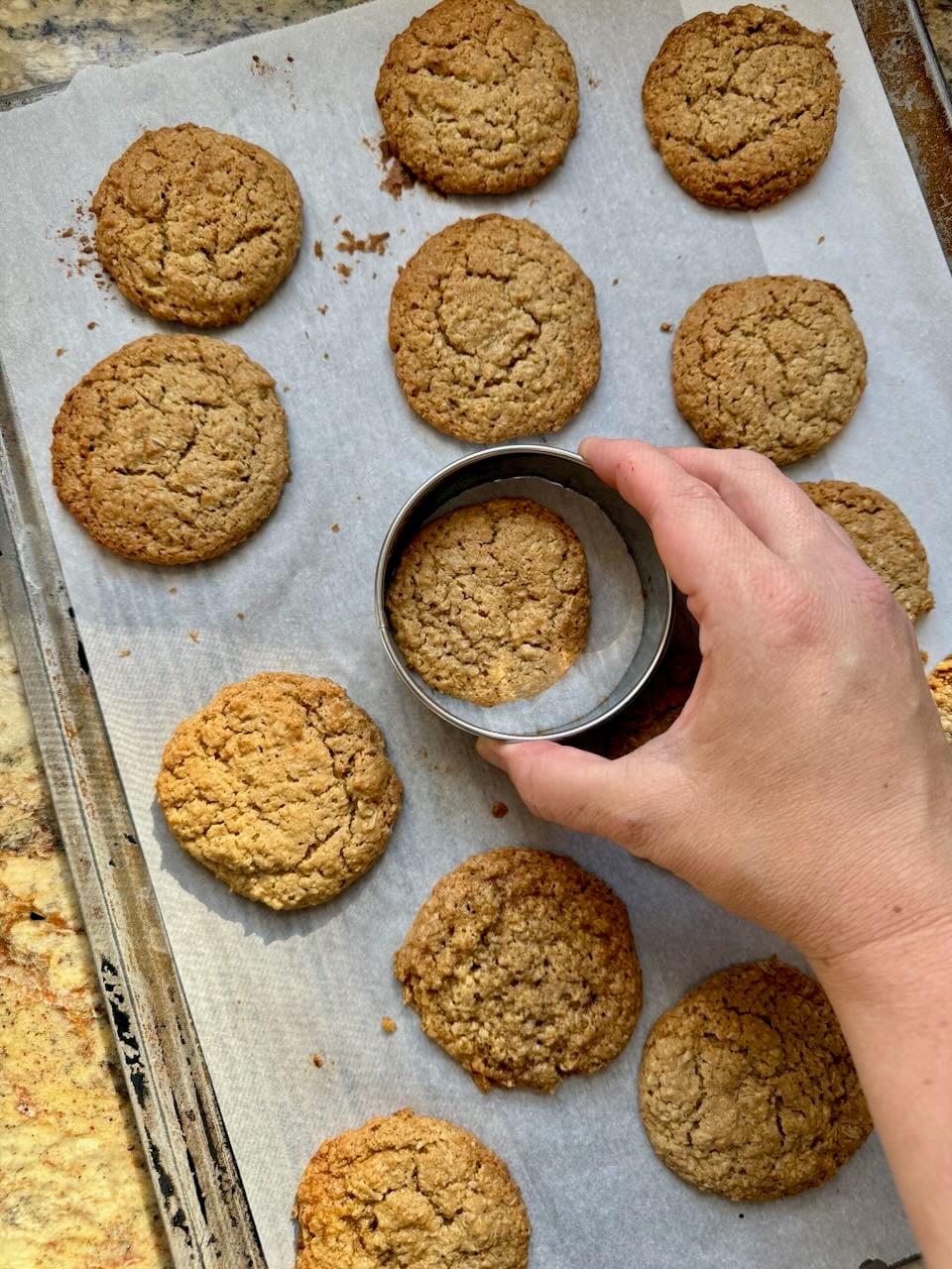 A hand holds a round biscuit cutter over a cookie, running it around the cookie to create uniform smooth round edges.