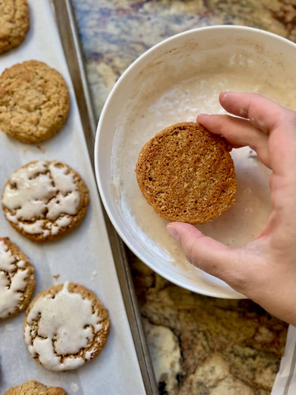 A cookie is held upside down in a bowl of icing alongside a cookie sheet containing iced cookies.