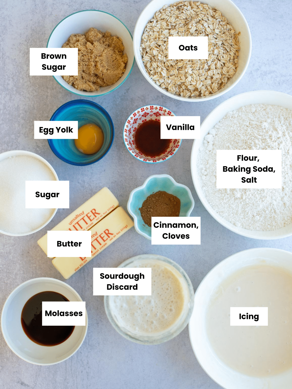 Overhead view of labeled baking ingredients for iced oatmeal sourdough cookies, including oats, sourdough discard, butter, molasses, brown sugar, and spices arranged neatly in bowls.