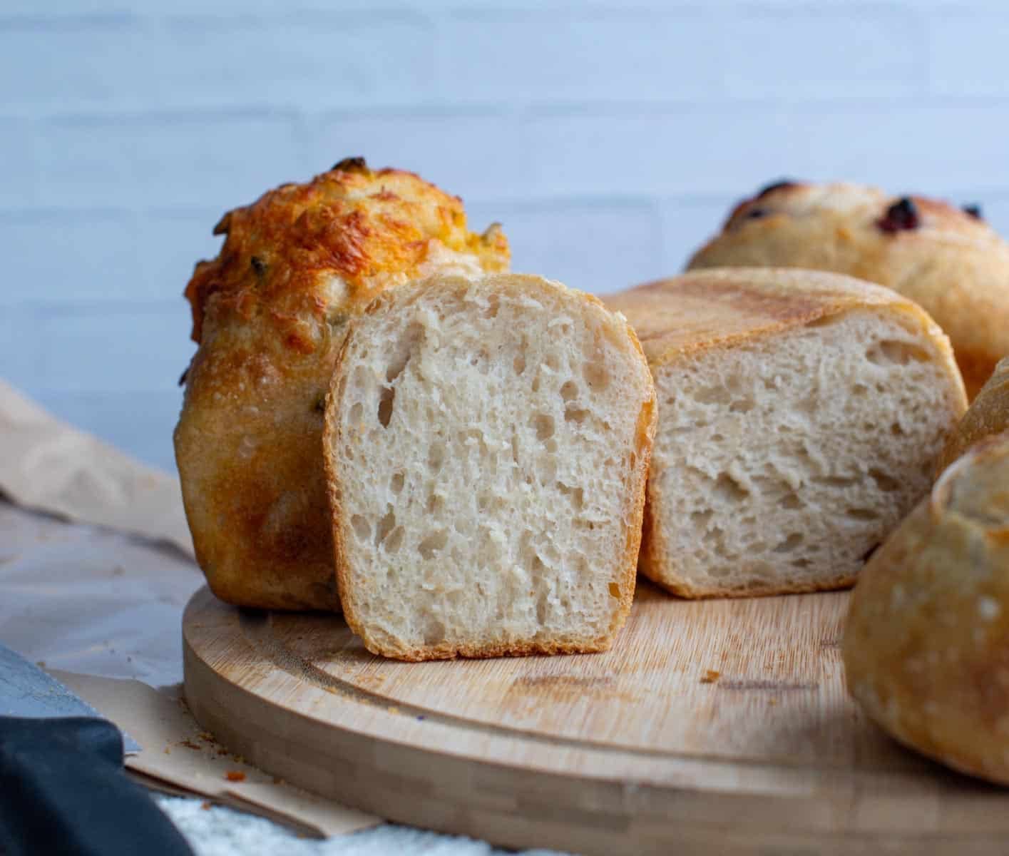 A sliced mini sourdough loaf reveals its soft, airy crumb beside several golden, rustic loaves on a wooden board.