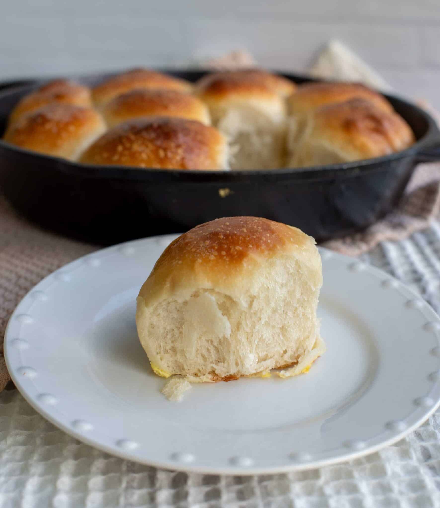 A single soft sourdough potato roll sits on a white plate, showing its fluffy, tender interior, with a cast iron skillet of golden brown rolls blurred in the background.
