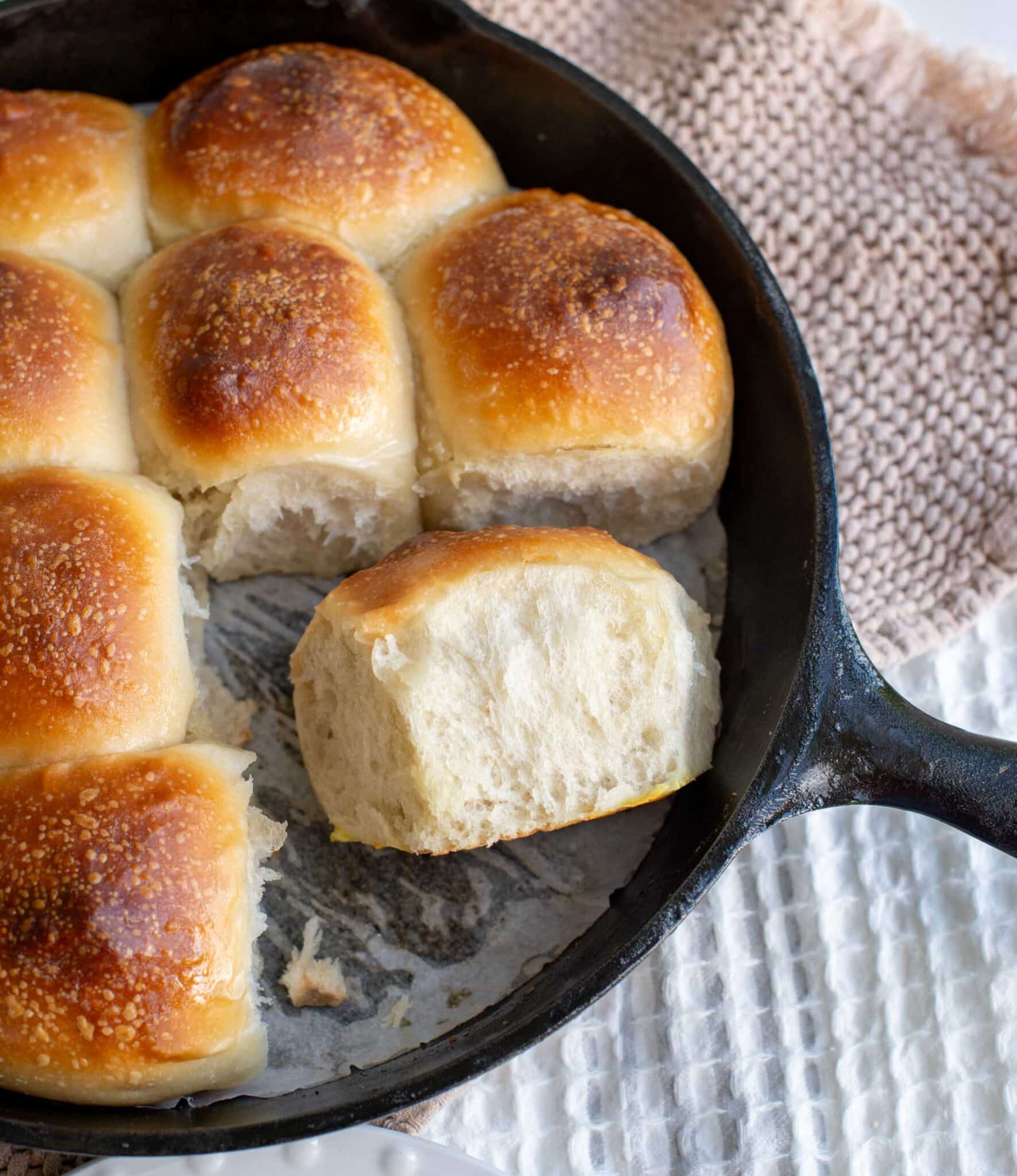 Golden brown soft sourdough potato rolls baked in a cast iron skillet, with one roll pulled apart to reveal a fluffy, tender interior.