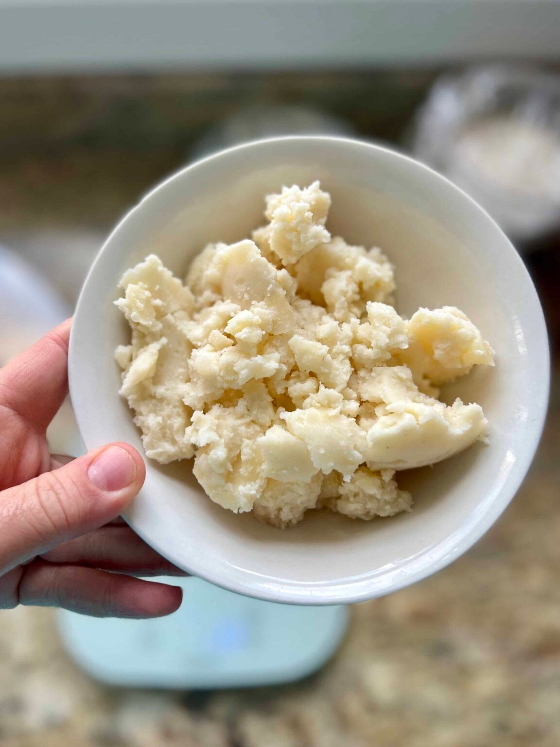 Mashed potatoes are in a white bowl held over a kitchen scale.