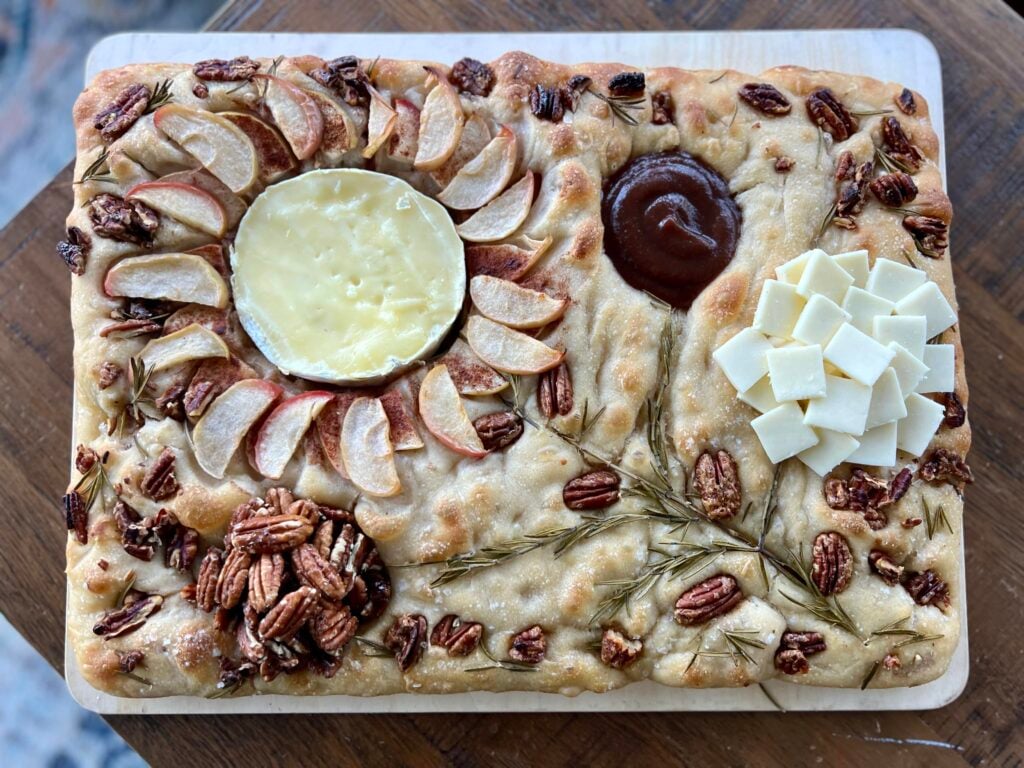 A focaccia charcuterie board is a large rectangle of focaccia containing baked in apple slices and pecans with other cheeses creating a flower pattern across the surface.