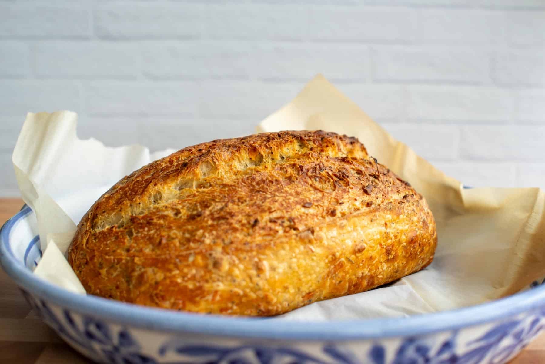 Golden-brown Italian herbs and cheese sourdough bread resting in a parchment-lined baking dish, with a crisp crust and rustic texture against a light brick background.
