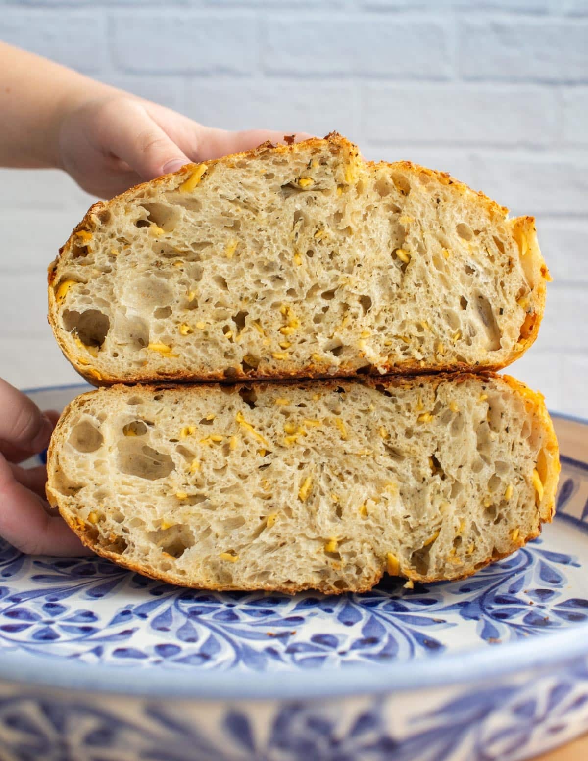 Sliced Italian herbs and cheese sourdough bread showing a soft, airy crumb with melted cheese pieces throughout, held above a patterned plate against a light background.