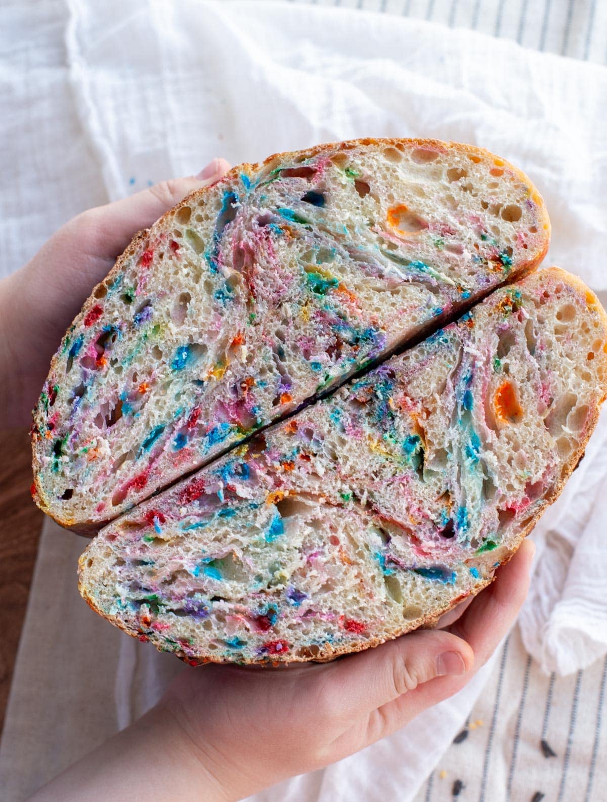 Hands holding a sliced Funfetti sourdough bread loaf, showing a soft, airy crumb filled with colorful rainbow sprinkles against a light fabric background.