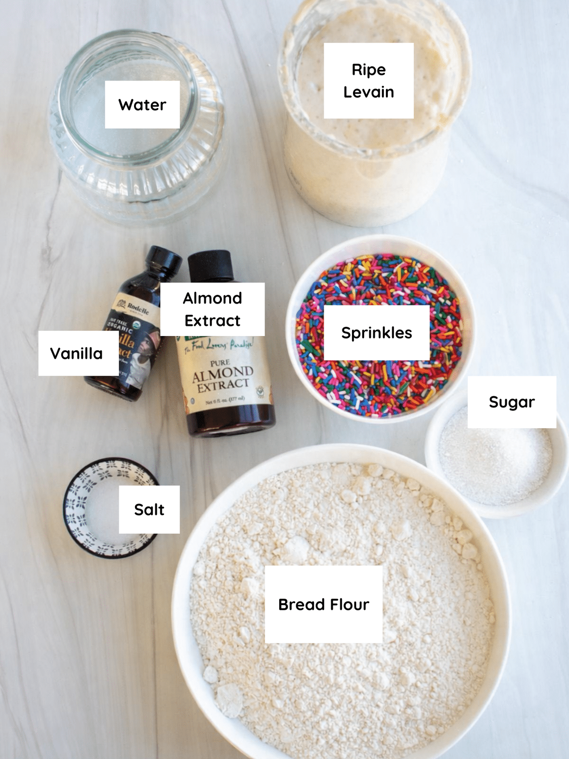 Labeled ingredients for Funfetti sourdough bread arranged on a light surface, including bread flour, ripe levain, water, sugar, salt, vanilla extract, almond extract, and rainbow sprinkles.
