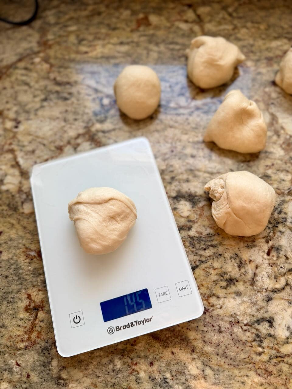 A ball of dough sits on a kitchen scale next to several other balls of dough.