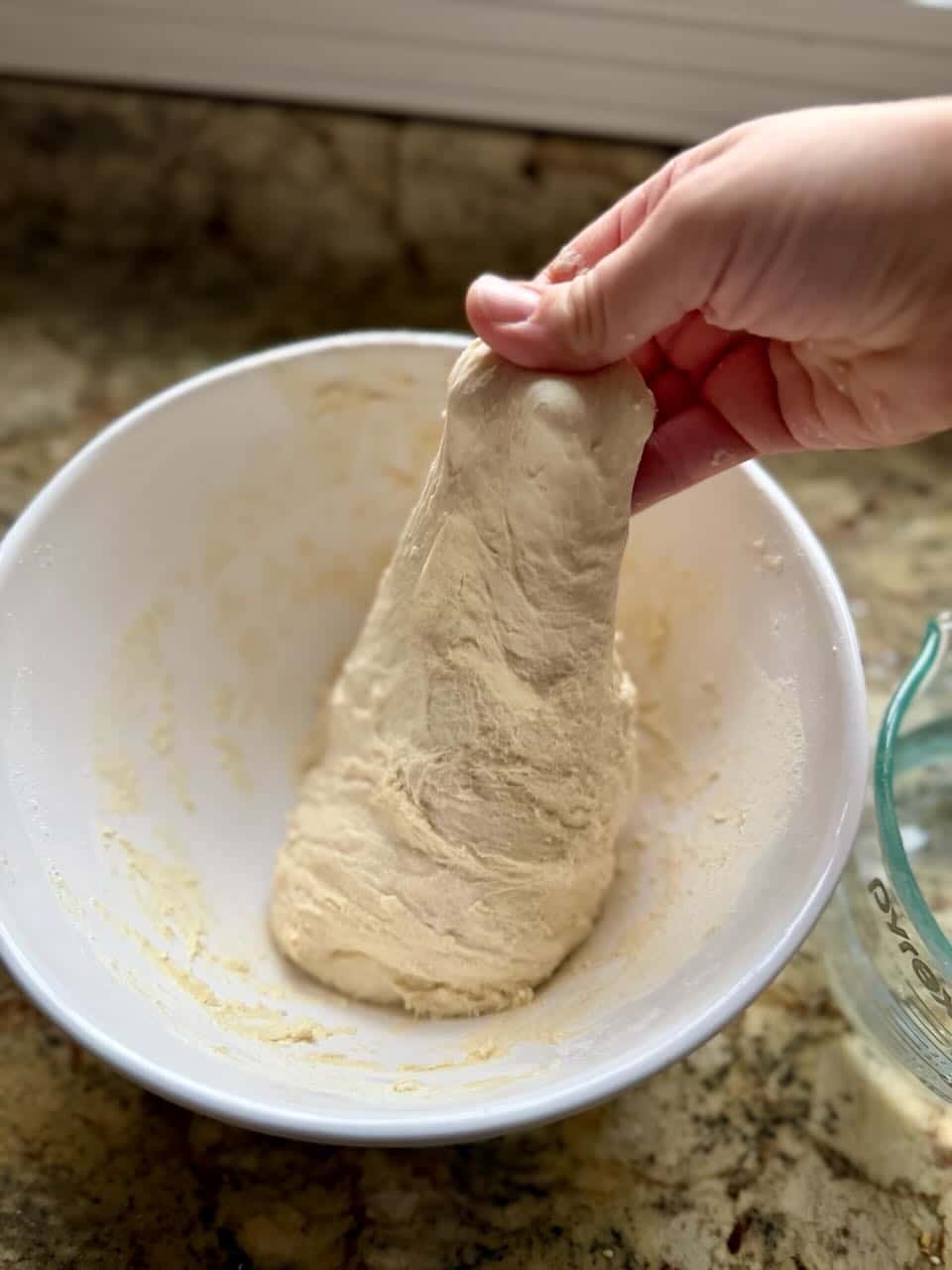 A hand lifts dough upwards out of a mixing bowl.