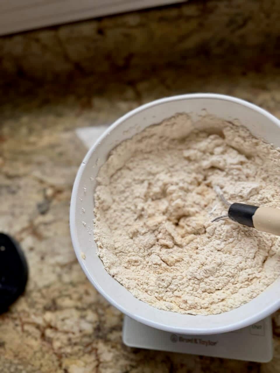 Flour is added to a mixing bowl that sits on a kitchen scale and has a dough whisk sitting in the bowl.