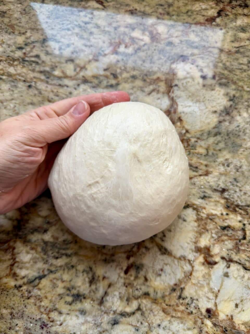 A hand touches the side of a round ball of dough on a kitchen counter.