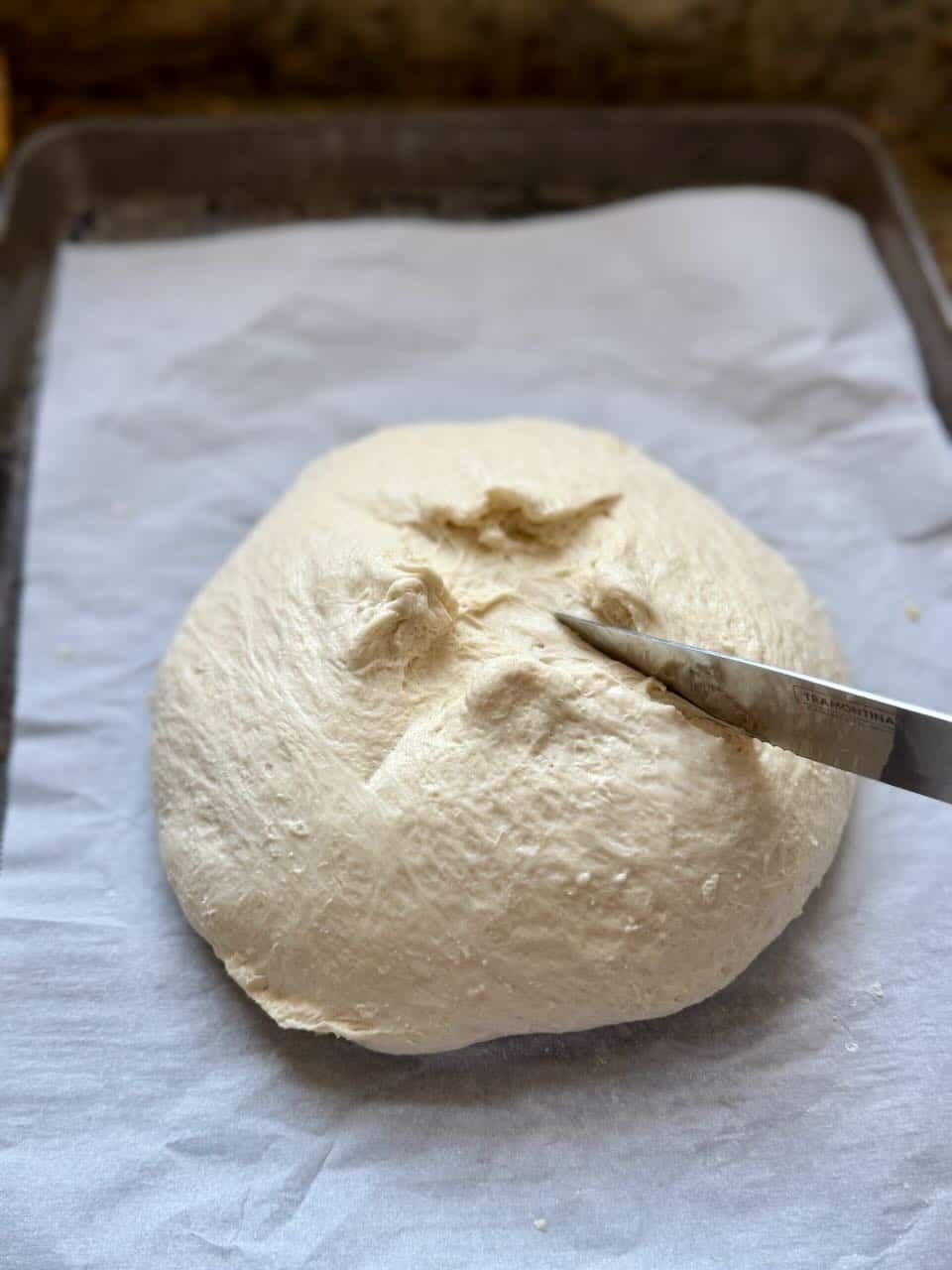 A knife slashes a cross score into a ball of dough sitting on parchment paper.