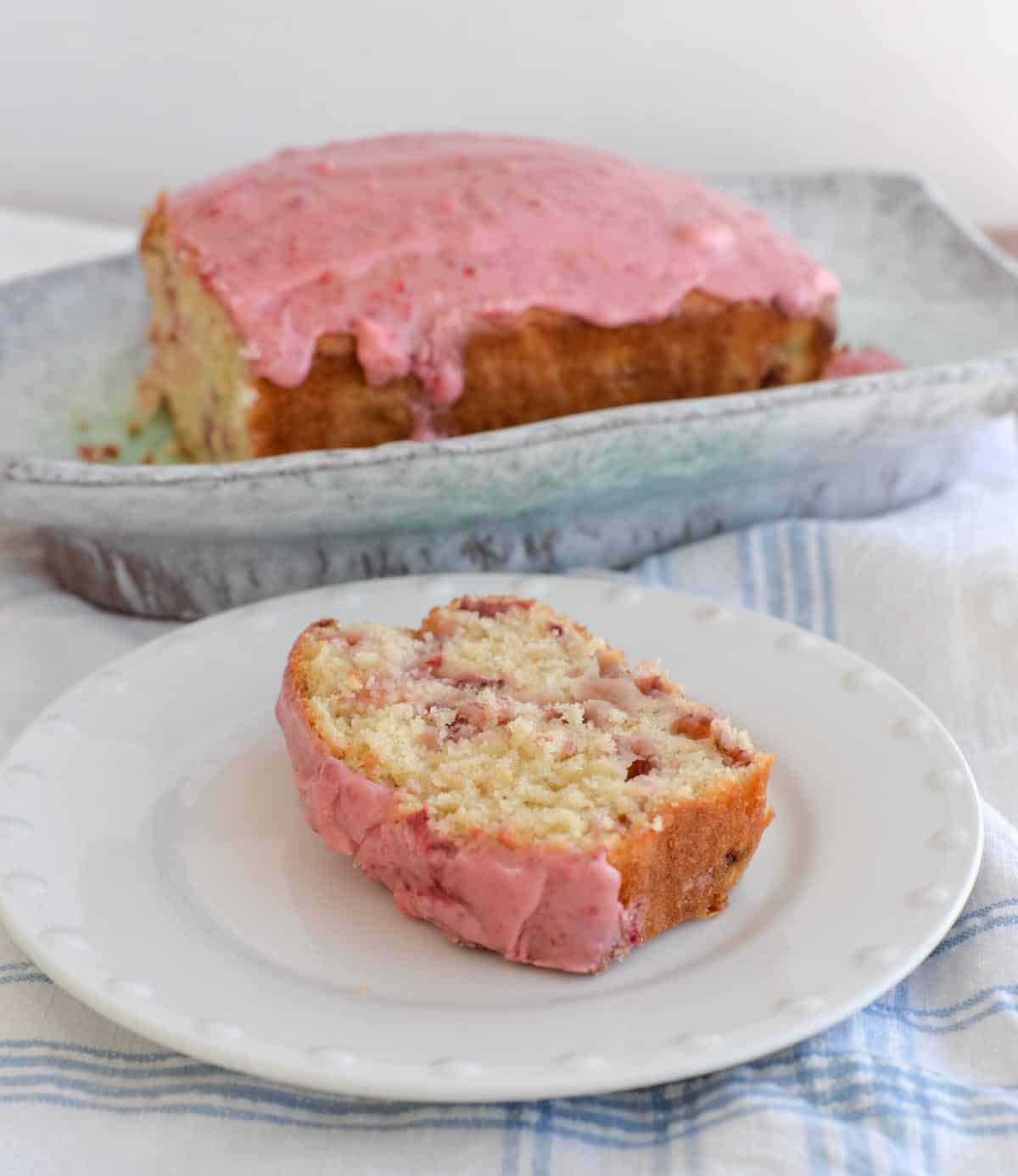 Slice of sourdough discard strawberry bread topped with pink strawberry glaze on a white plate, with the glazed loaf in the background.