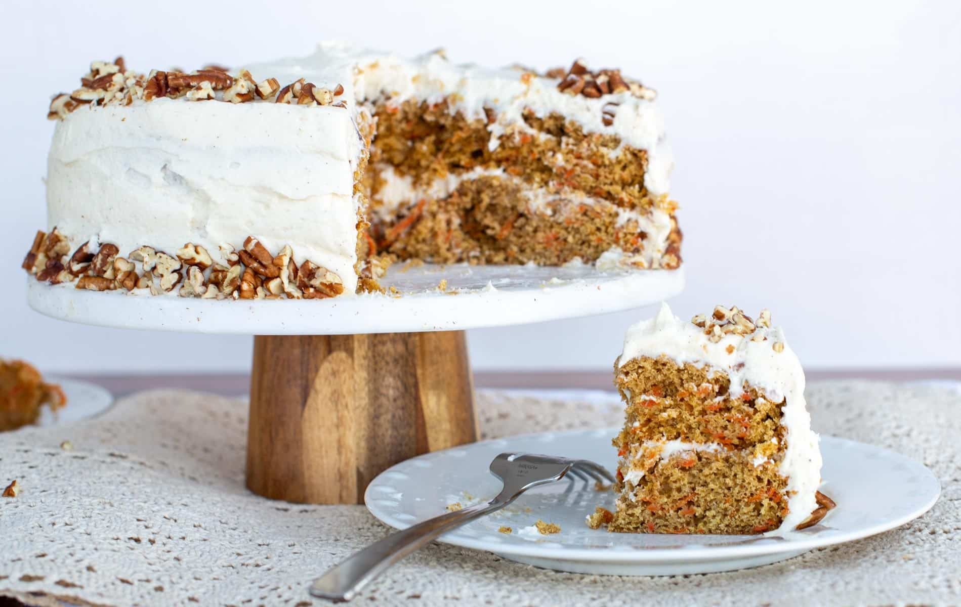 Layered sourdough carrot cake with cream cheese frosting and chopped pecans on a cake stand, with a sliced piece served on a plate in the foreground.