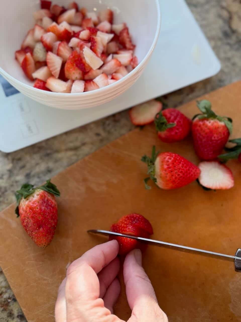 A hand slices fresh strawberries on a cutting board beside a bowl of diced strawberries resting on a kitchen scale.