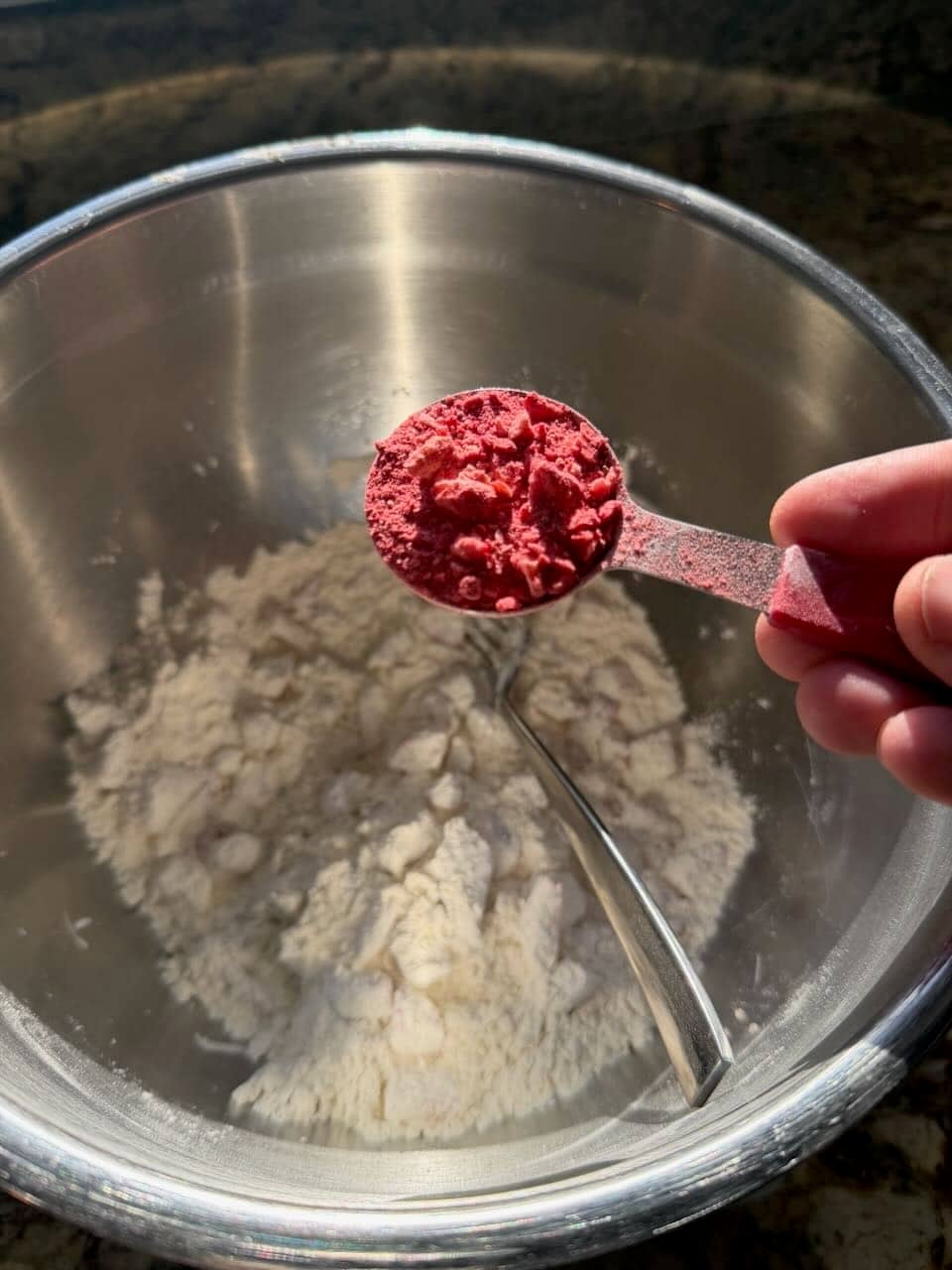 A measuring spoon filled with crushed freeze-dried strawberries is held over a bowl of flour in a metal mixing bowl.