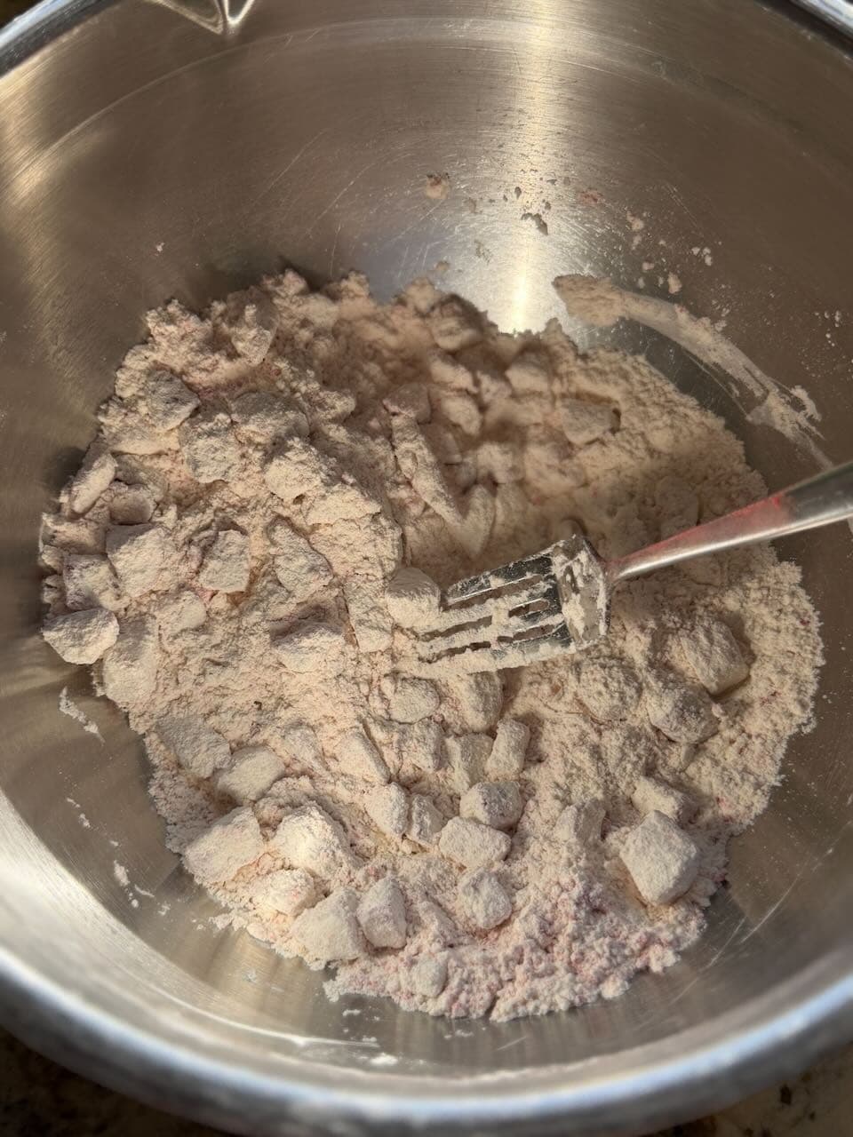 Flour mixed with freeze-dried strawberry pieces sits in a metal bowl with a fork partially stirring the dry ingredients.