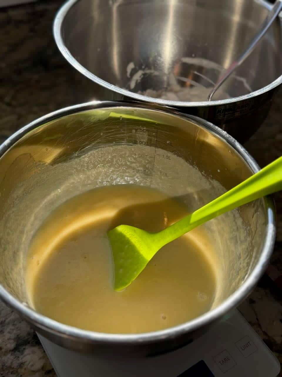 A bowl of pale yellow wet ingredients is being mixed with a green spatula on a kitchen scale.