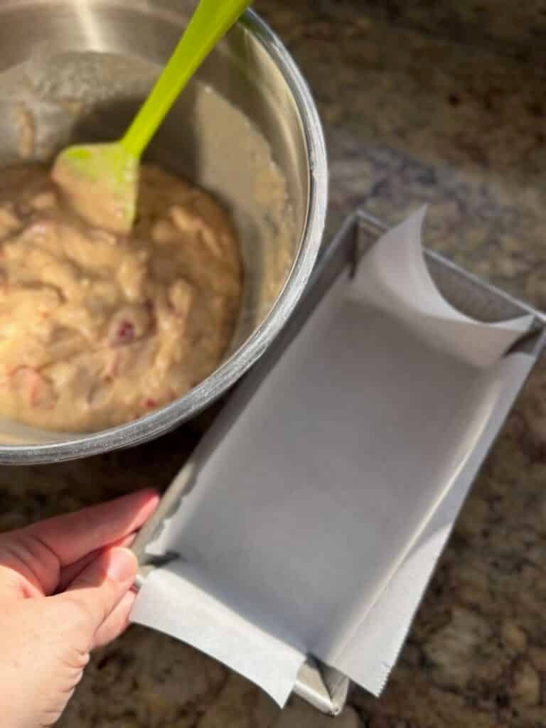 Strawberry bread batter is stirred in a bowl next to a parchment-lined loaf pan ready for filling.