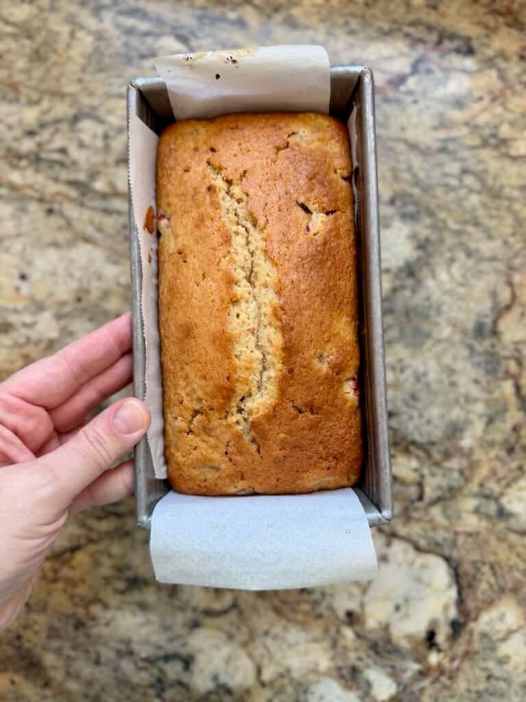 A freshly baked strawberry loaf sits in a parchment-lined pan, golden brown with a crack down the center.