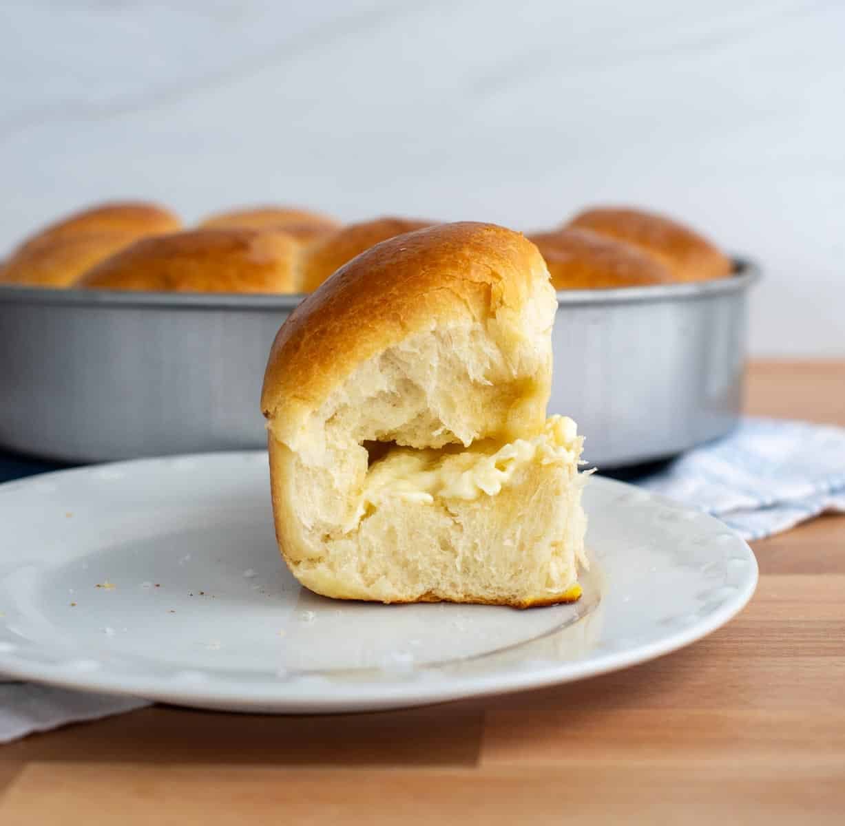Soft sourdough brioche roll split open and spread with butter, showing the fluffy interior with a pan of golden rolls in the background.